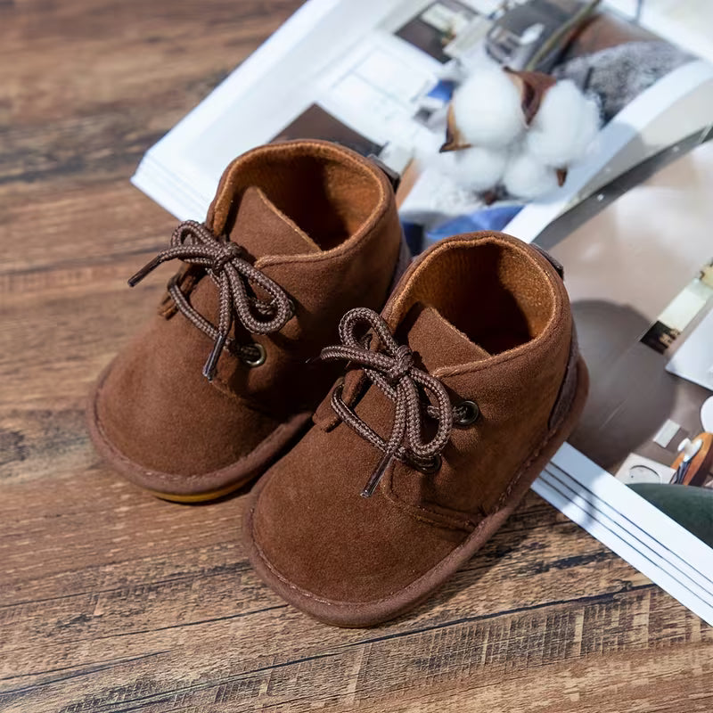 Pair of brown suede baby boots on a wooden surface with a magazine underneath.