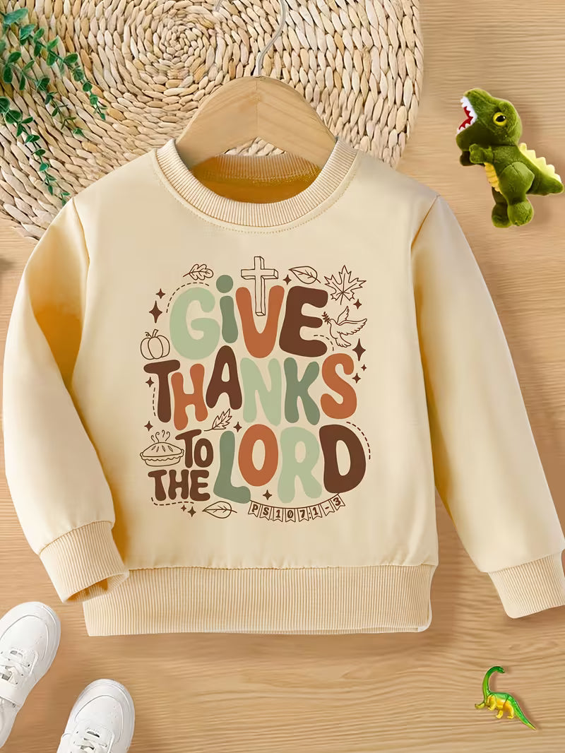 Beige children's sweatshirt with 'Give Thanks to the Lord' text on a wooden surface.